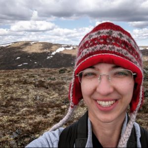 Headshot of Christin Swearingen, smiling and wearing a red hat on a tundra landscape